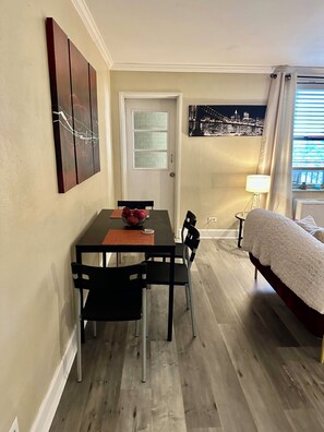A compact room with a dining table, chairs, a bed, and a cityscape painting on the wall.