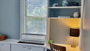 Habitación con escritorio integrado, estanterías, una ventana con persianas, una silla y una lámpara.