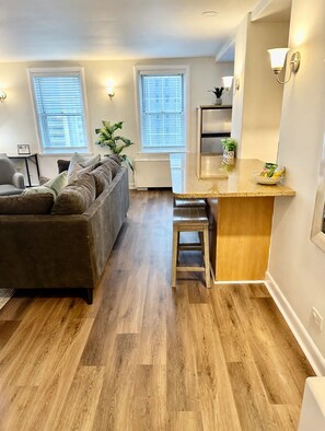 A modern living room with a grey sofa, a wooden coffee table, and a kitchen area with a wooden countertop and bar stools. - FIFA 2026 Walk to Stadium | 1BR Unit 12K ATL (Atlanta)
