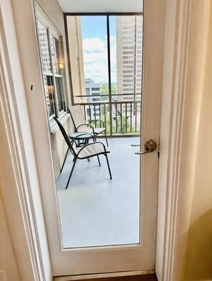 A door to a balcony with two chairs and a table, overlooking a cityscape.