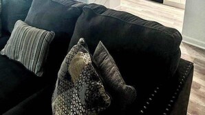 Una sala de estar moderna con un sofá negro y cojines estampados, que conduce a una cocina con armarios blancos y electrodomésticos de acero inoxidable.