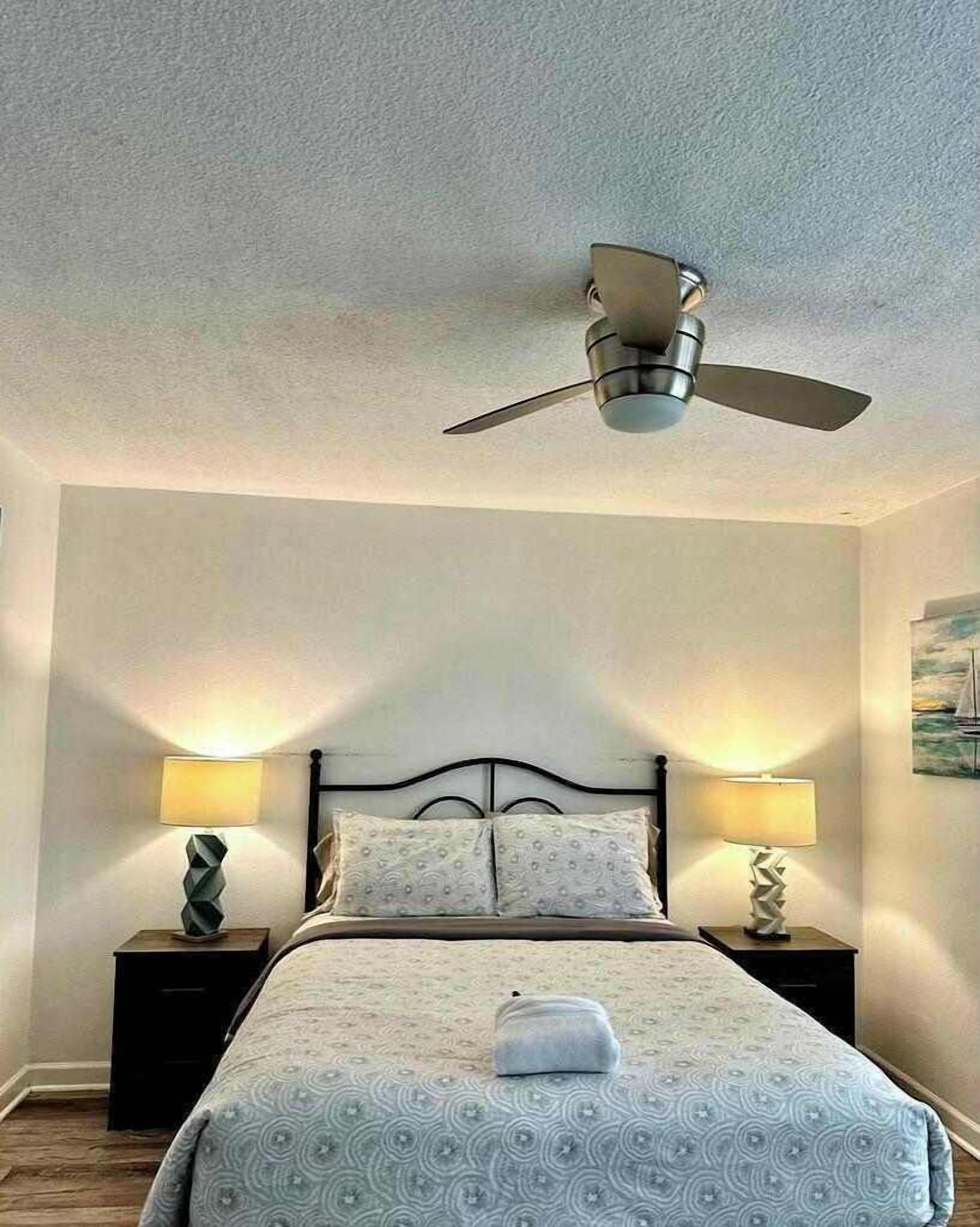 A bedroom with a bed, two bedside tables with lamps, a ceiling fan, and a painting on the wall.