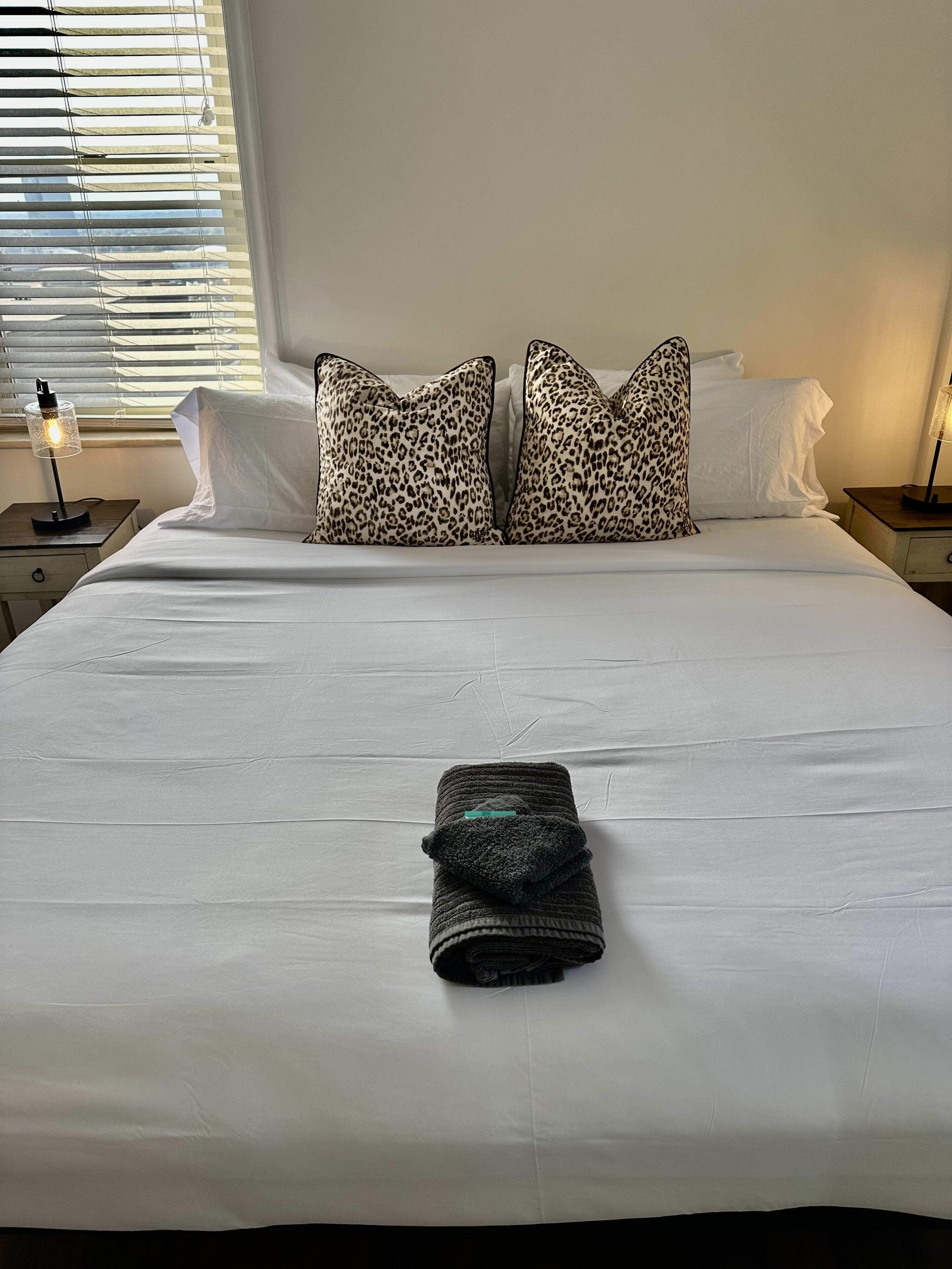 A neatly made bed with white sheets, two leopard print pillows, and a folded dark towel on the bed.