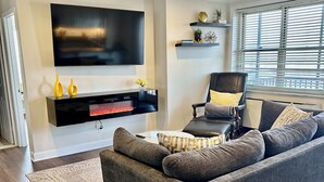 A modern living room with a grey sofa, a flat-screen TV mounted on the wall, a black entertainment unit, and a window with blinds. - FIFA 2026 Walk to Stadium | 1BR Unit 24B ATL (Atlanta)