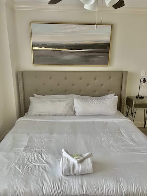 A neatly made bed with a white duvet, a folded towel, and a painting of a landscape above it.