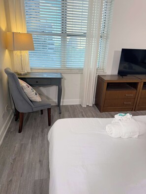 A hotel room with a bed, a desk with a lamp, a television, and a window with blinds. - Comfortable Downtown 1BR | Walk to the Stadium (Atlanta)