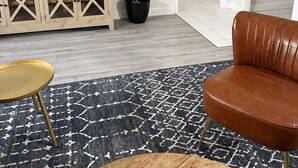 Una sala de estar con televisor, una planta en maceta, un sofá de cuero marrón, una mesita redonda y un tapete estampado.