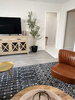 Una sala de estar con televisor, una planta en maceta, un sofá de cuero marrón, una mesita redonda y un tapete estampado.
