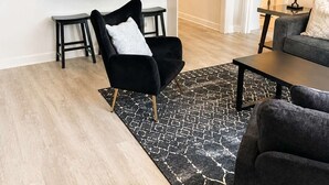 A modern living room with a black sofa, a black armchair, a coffee table, a patterned rug, and a kitchen area with a counter and stools. - FIFA 2026 Walk to Stadium | 1BR Unit 4E ATL (Atlanta)