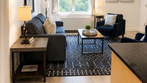 A modern living room with a sofa, two armchairs, a coffee table, and a small side table. There is a window with curtains, a ceiling fan, and artwork on the wall. - FIFA 2026 Walk to Stadium | 1BR Unit 4E ATL (Atlanta)