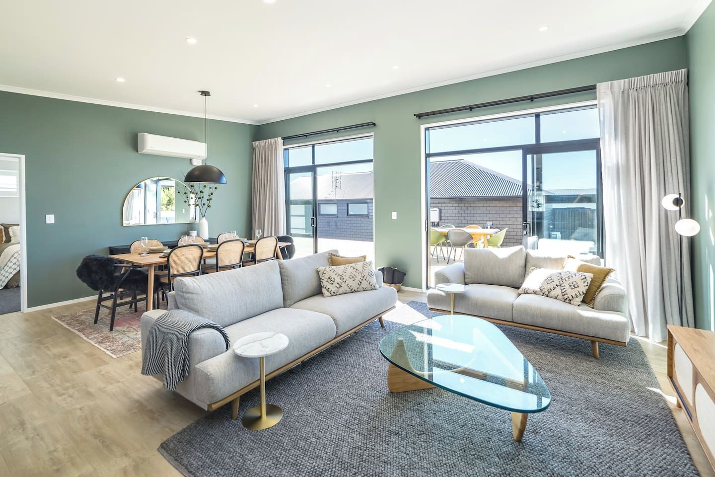 A modern living room with a sofa, armchair, and coffee table. There is a dining area with chairs and a table. Large windows provide natural light.