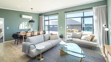 A modern living room with a sofa, armchair, and coffee table. There is a dining area with chairs and a table. Large windows provide natural light.