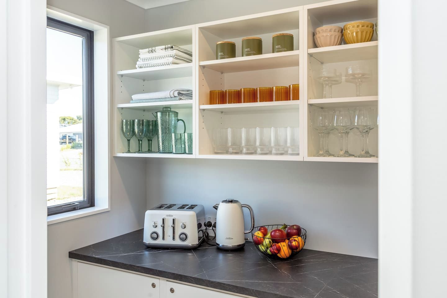 A kitchen with white cabinets, a window, a countertop with a toaster, a kettle, and a bowl of fruit.
