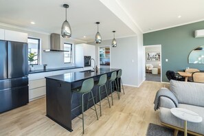 A modern kitchen with a central island, bar stools, pendant lighting, and a dining area with a sofa and table. - Sunset On Syrah, Martinborough (Martinborough)