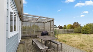 Une terrasse en bois avec une table et des bancs, un espace salon couvert et une vue sur le jardin arrière.