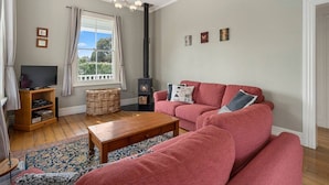 A living room with a red sofa, a wooden coffee table, a television on a stand, a fireplace, and a large window with curtains. - Applecross Cottage | Cozy Stay, Walk to Martinborough Square (Martinborough)
