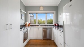 A modern kitchen with white cabinets, a stainless steel stove, a dishwasher, and a large window with a view of greenery. - Applecross Cottage | Cozy Stay, Walk to Martinborough Square (Martinborough)