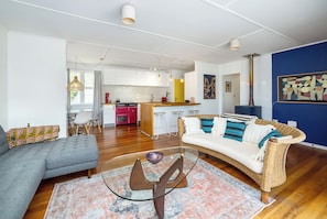 A modern living room with a grey sofa, a wicker sofa with blue cushions, a glass coffee table, and a kitchen in the background.