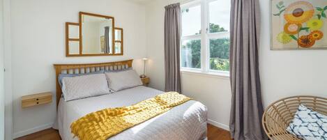 A bedroom with a bed, a bedside table, a chair, a window with curtains, and a painting on the wall.