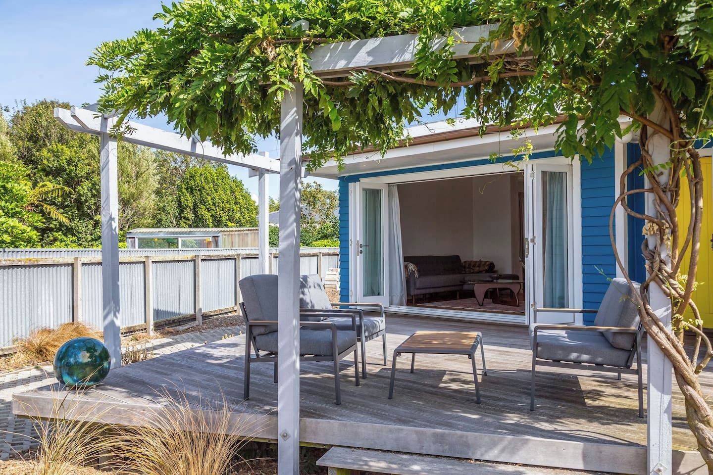 A covered outdoor seating area with a pergola, a sliding glass door leading to a room with a couch and a table, and a wooden deck.