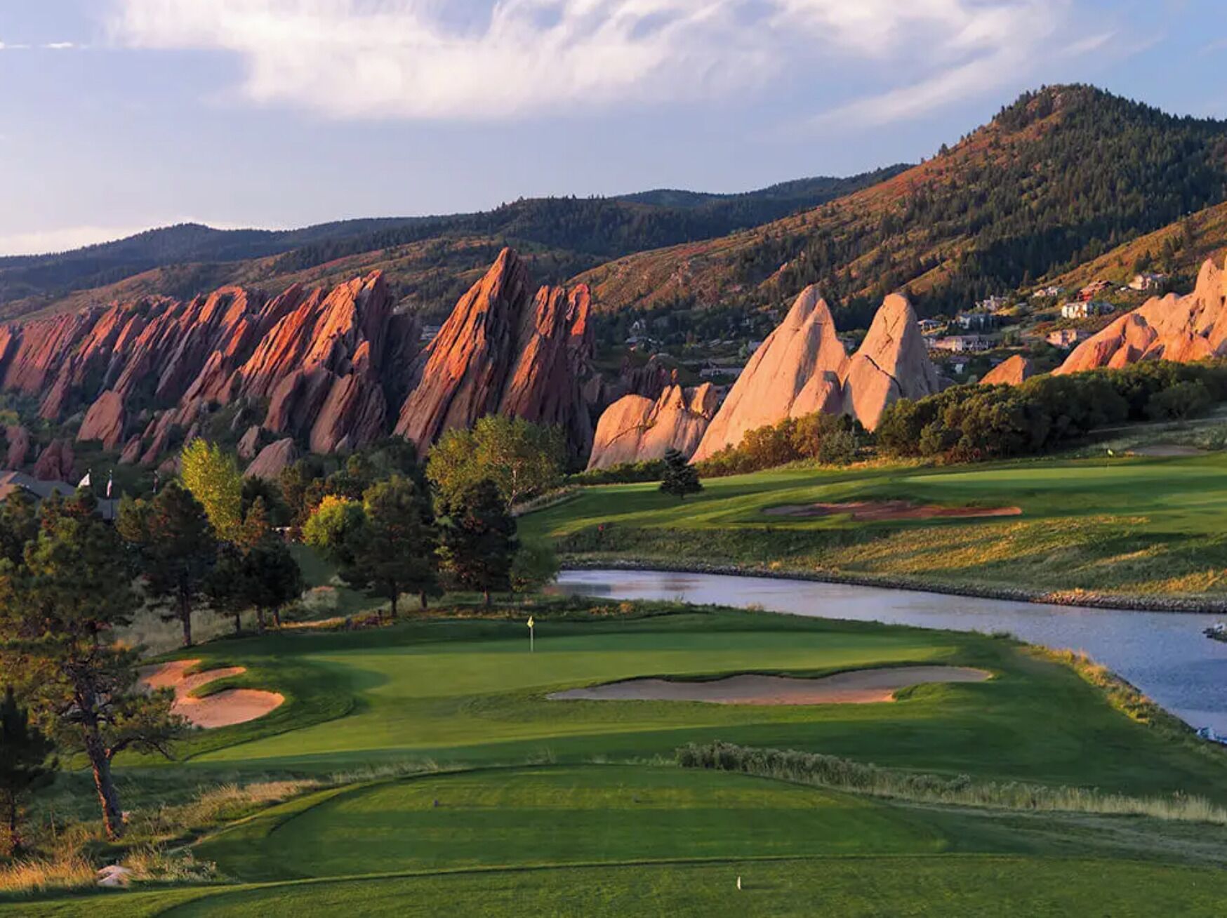 Un campo de golf con un obstáculo acuático, árboles y formaciones rocosas rojas al fondo.