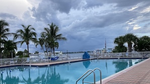 Une piscine dont le bord est revêtu de carreaux, entourée de chaises longues et de palmiers, et offrant une vue sur la mer et un ciel nuageux.