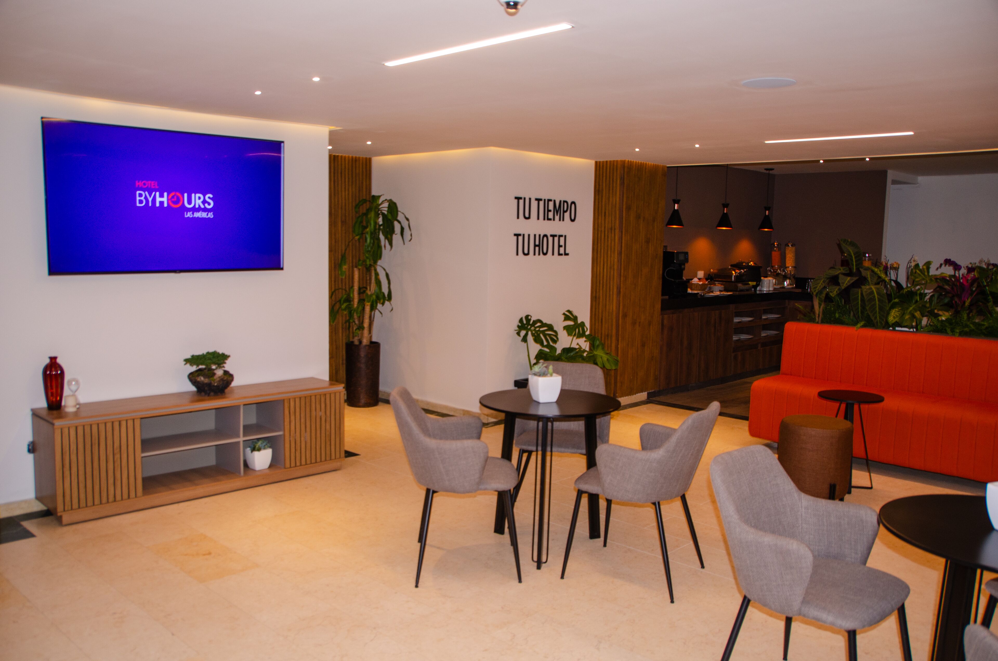 En moderne hotellobby med et fladskærms-tv, der viser 'Hotel ByHours', et spiseområde med grå stole, en rød sofa og et tekøkken.
