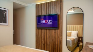 Une chambre d’hôtel avec un lit, un mur recouvert de panneaux en bois, un téléviseur affichant le logo « Hotel ByHours Las Americas », et un miroir qui reflète le lit et une table de chevet.