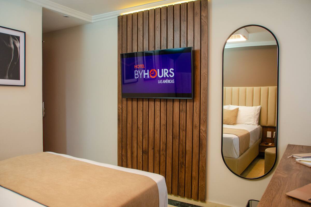 Ein Hotelzimmer mit einem Bett, einer Wand aus Holzpaneelen, einem Fernseher, der 'Hotel ByHours Las Americas' anzeigt, und einem Spiegel, der das Bett und einen Nachttisch reflektiert.