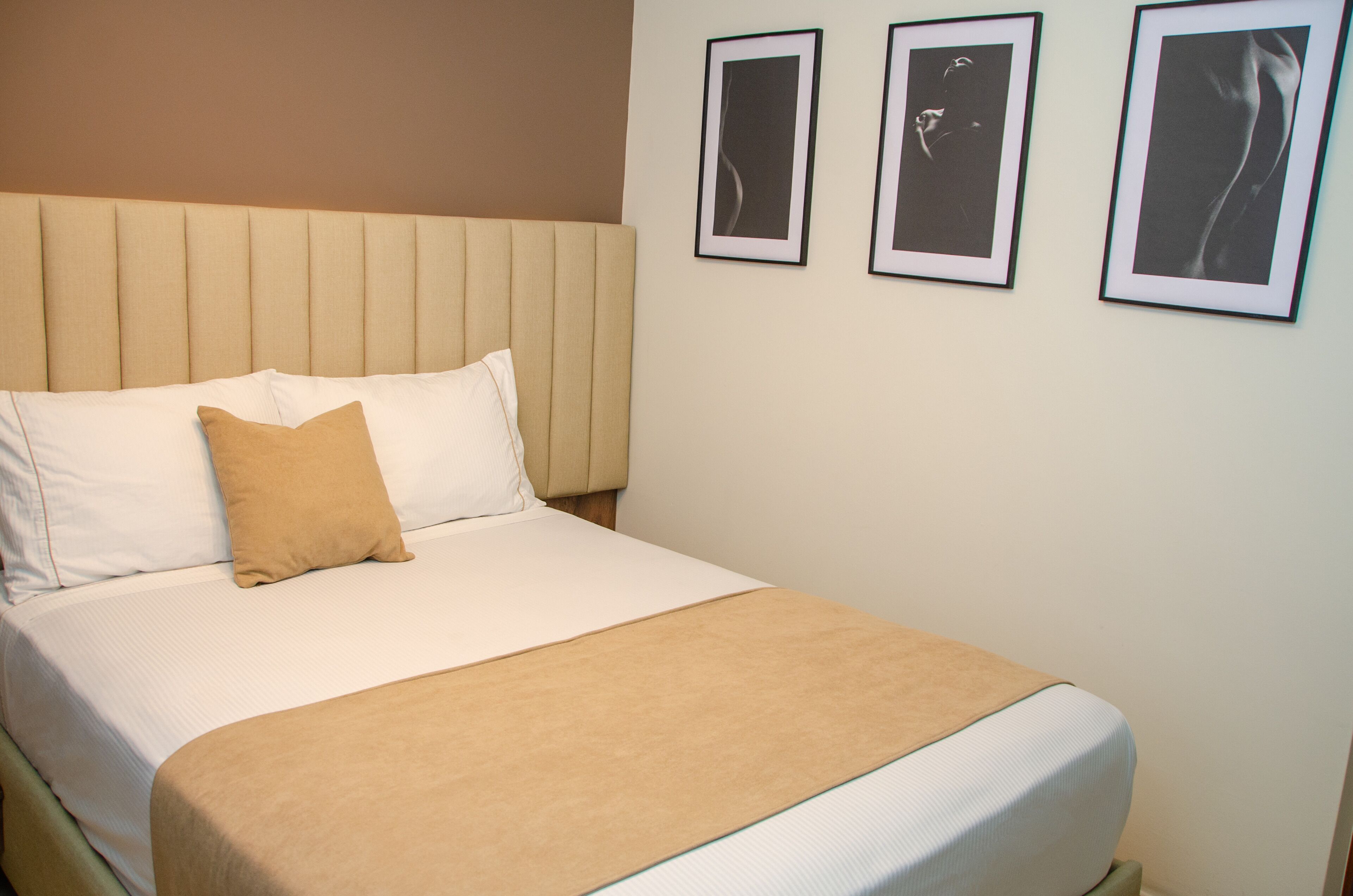 Una cama bien hecha con un cabecero color beige, ropa de cama blanca y una manta color arena. Hay tres cuadros enmarcados en la pared sobre la cama.