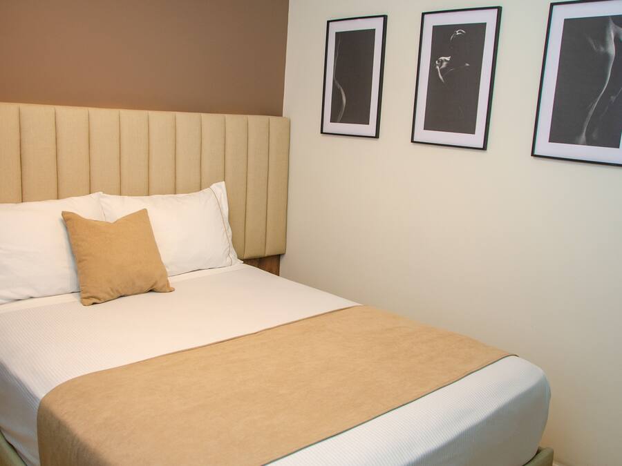 Una cama bien hecha con un cabecero color beige, ropa de cama blanca y una manta color arena. Hay tres cuadros enmarcados en la pared sobre la cama.