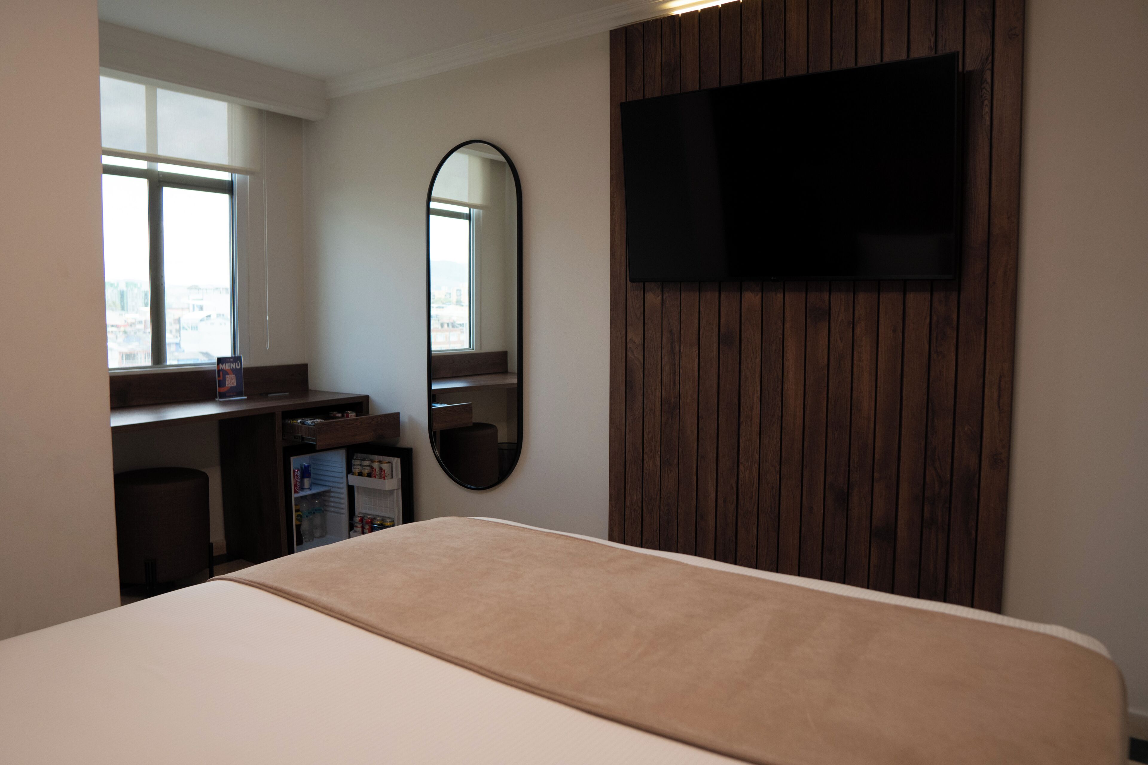 Habitación de hotel con cama, televisión montada en una pared de paneles de madera, espejo, minibar y ventana con vistas a edificios.