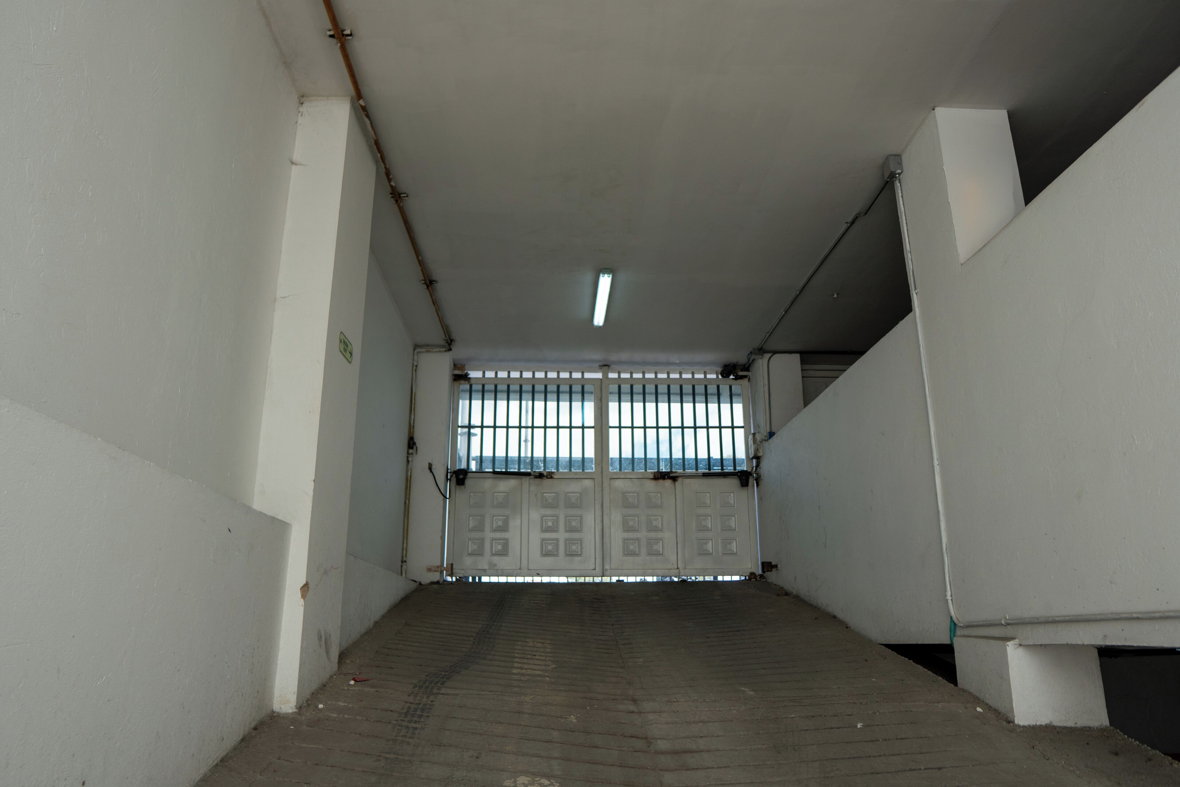 Un pasillo con entrada protegida por barrotes, paredes de concreto y una lámpara fluorescente.