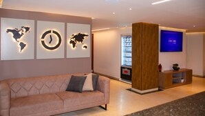 Un hall d’entrée moderne avec un canapé, une carte du monde accrochée au mur, une horloge, un distributeur automatique et un écran de télévision affichant le logo « BYHOURS ».