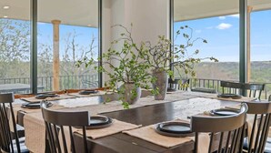A dining table set for six with a vase of green plants in the center, large windows on either side, and a view of the outdoors. - Ranch Retreat: Amenities Galore! (Leander)