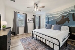 A bedroom with a bed, a ceiling fan, a desk with a lamp, a chair, a rug, and a mural on the wall. - Ranch Retreat: Amenities Galore! (Leander)