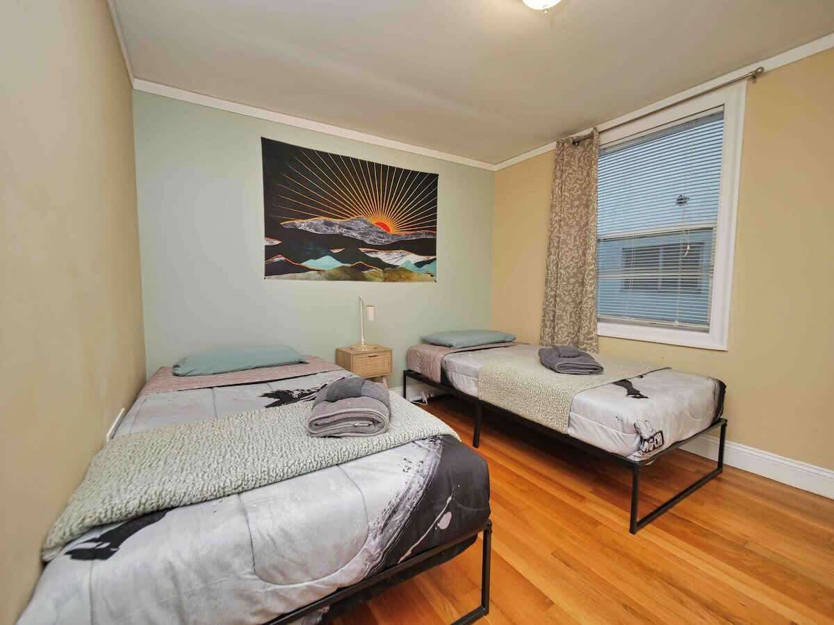 Una habitación con dos camas individuales, una mesita de noche, un cuadro colgado en la pared, una ventana con persianas y suelo de madera.