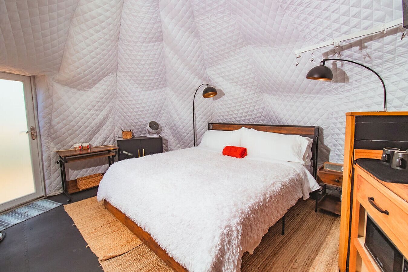 A cozy bedroom with a large bed, a wooden dresser, a nightstand, a lamp, and a quilted ceiling.