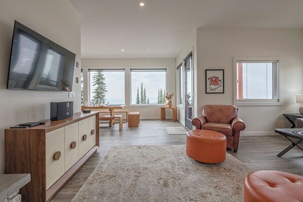 Uma sala de estar moderna com uma televisão grande, mobiliário de madeira e vista para árvores através de grandes janelas.