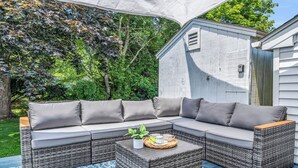 A covered outdoor seating area with a wicker sofa and a coffee table, surrounded by greenery. - Great Hammcock Getaway, cozy home, hot tub, fire pit, beaches nearby. (Old Saybrook)