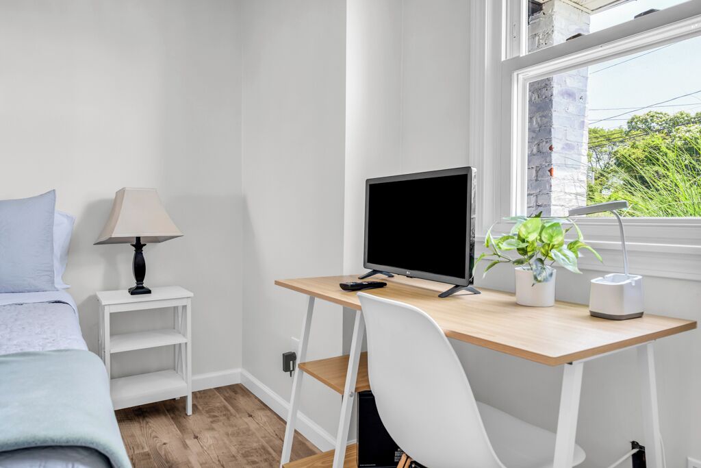 Et soveværelse med seng, skrivebord med computer, lampe og vindue med plante.