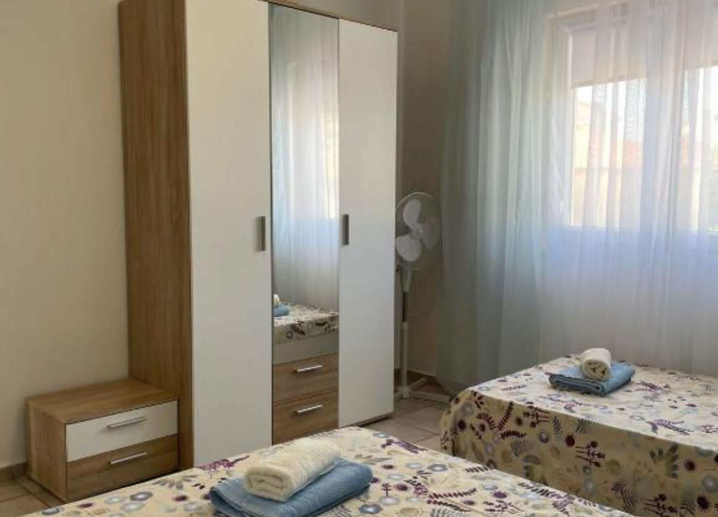A bedroom with a wardrobe, two beds, a bedside table, and a window with curtains.