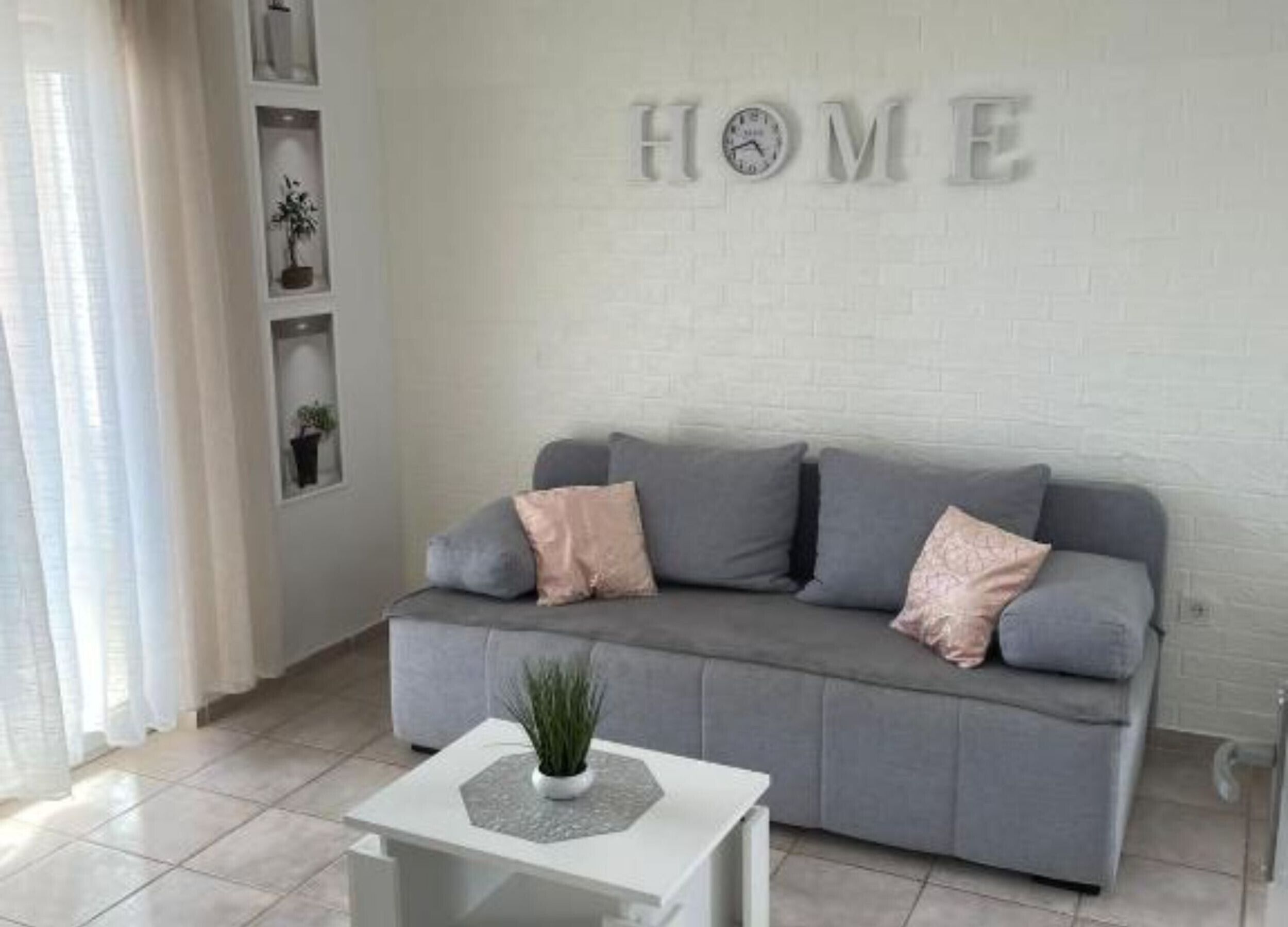 A modern living room with a grey sofa, a small white table with a plant, and a wall-mounted clock. Shelves with decorative items are visible.