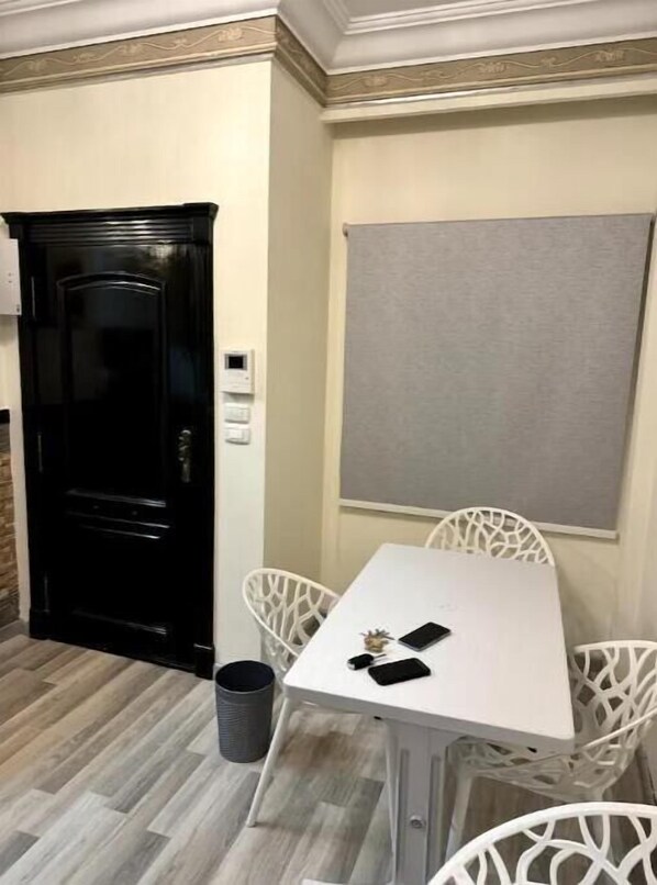 A compact room with a white table, four white chairs, a black door, a small trash bin, and a grey wall panel. - 820 · 2BR Townhouse | 2 Queen Beds | Backyard (Sheikh Zayed City)