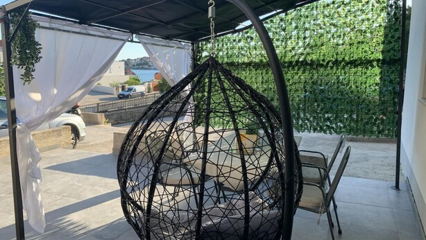 A hanging chair under a covered patio area with a view of a green hedge and a body of water in the background. - Apartments J&M 2 - One Bedroom Apartment with Terrace (Rogoznica)