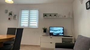 A modern living room with a dining area, featuring a TV, white shelves with plants, and a window with blinds. - Apartments J&M 2 - One Bedroom Apartment with Terrace (Rogoznica)