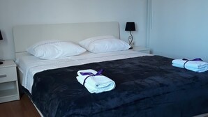 Una cama bien hecha con una manta azul oscuro y dos toallas, un cabecero blanco, una lámpara de noche y un aplique de pared.