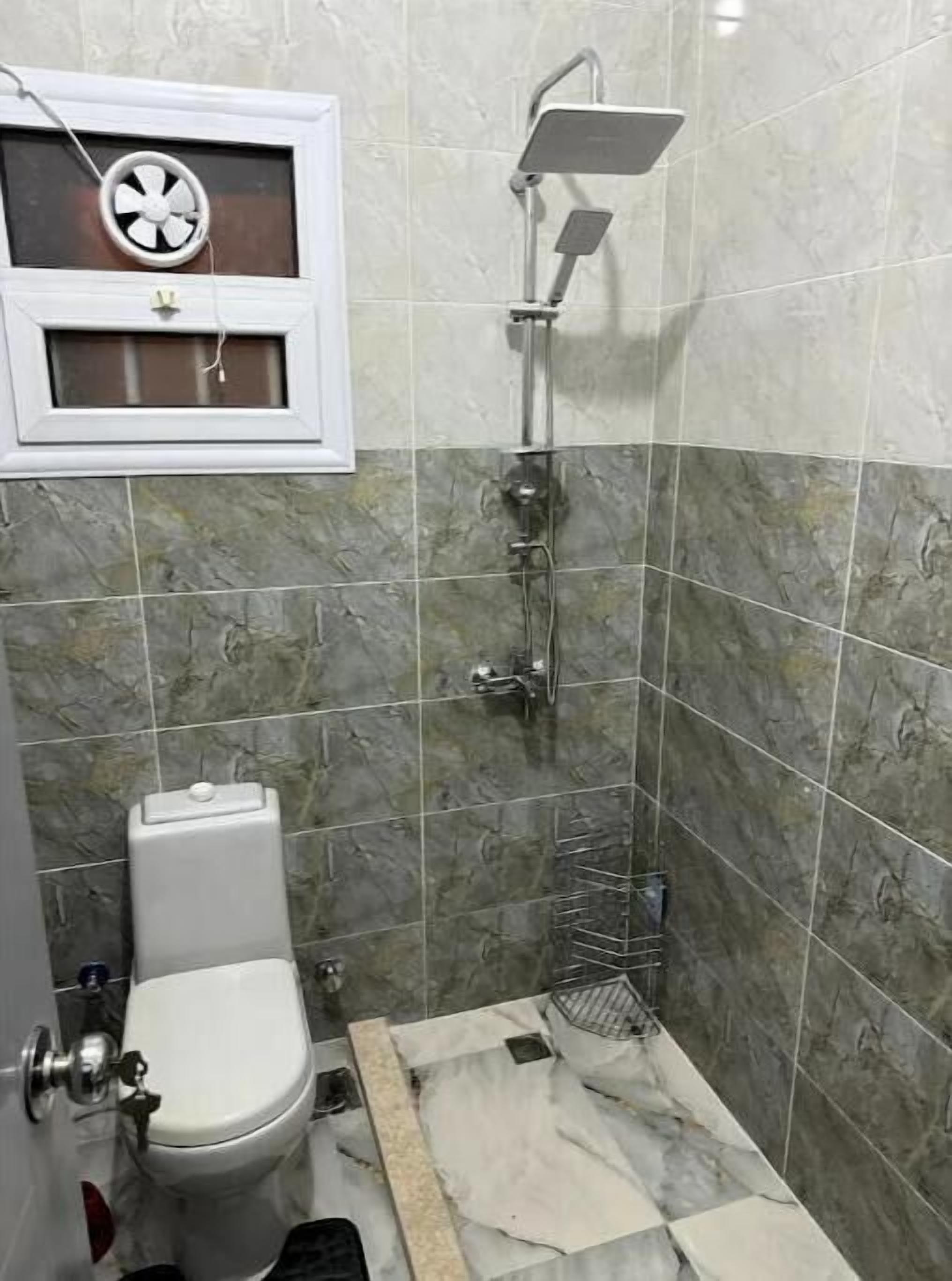 Une petite salle de bain comprenant des toilettes, une douche et une petite fenêtre avec un ventilateur.