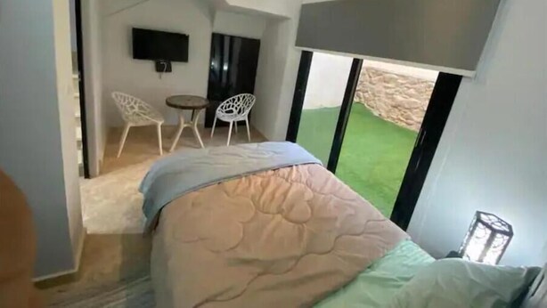 A bedroom with a bed, a TV, a small table, two chairs, and a view of a green outdoor area through large windows. - 819 · 2BR Townhouse | Garden | Sleeps 5 (Sheikh Zayed City)