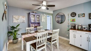 Uma área de refeições com uma mesa posta para quatro pessoas, cadeiras brancas, um ventilador de teto e uma cozinha com armários e um espaço para café.